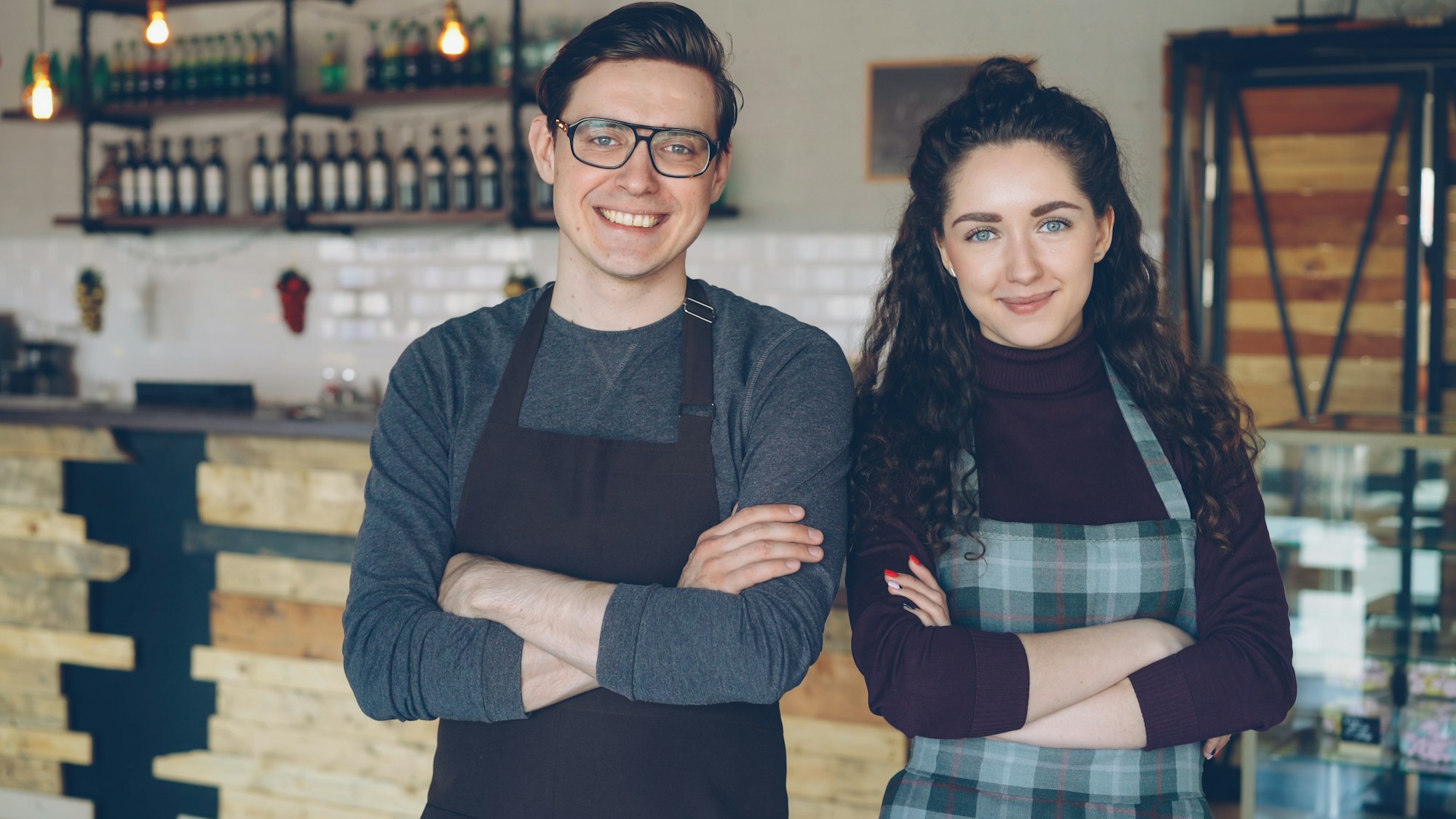 Two smiling cafe owners in aprons, arms crossed, in front of their counter